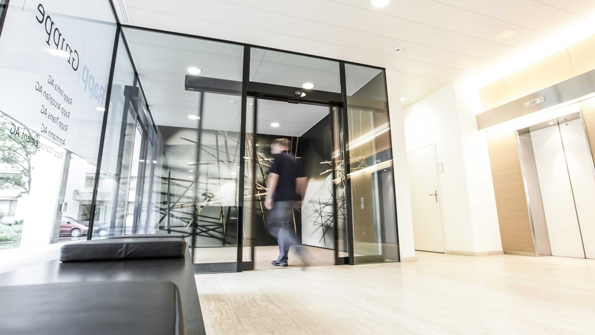 Man entering a glass-walled corporate lobby with abstract wall art and bright lighting behind it, motion blurred as he walks through the door.